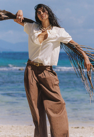a-woman-stands-on-a-beach-holding-a-palm-frond-wearing-a-loose-white-blouse-with-lace-trim-brown-wide-leg-pants-and-a-patterned-belt
