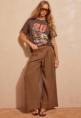 a-woman-modeling-a-dark-brown-graphic-t-shirt-and-matching-brown-wide-leg-pleated-trousers-styled-with-a-brown-belt-sunglasses-and-sandals-against-a-textured-beige-wall