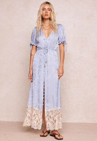 a-woman-modeling-a-light-blue-maxi-dress-with-short-puff-sleeves-a-v-neckline-and-a-lace-hem-standing-against-a-textured-beige-wall