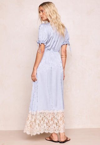 a-woman-modeling-a-light-blue-maxi-dress-with-short-puff-sleeves-side-tie-details-and-a-cream-lace-hem-shown-from-the-back-against-a-textured-beige-wall