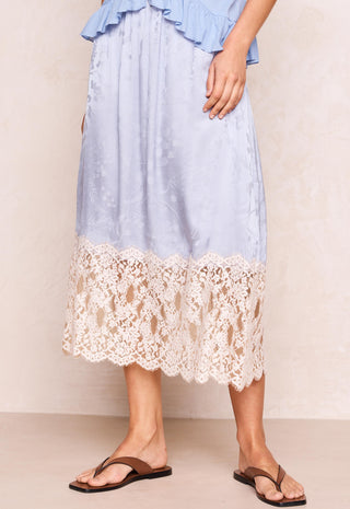 a-light-blue-midi-skirt-with-a-cream-lace-hem-shown-on-a-model-wearing-brown-thong-sandals-against-a-neutral-beige-background