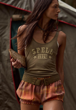 a-woman-wearing-a-vintage-style-olive-green-tank-top-with-spell-1976-printed-on-the-front-paired-with-plaid-shorts-standing-in-front-of-a-tent