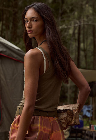 a-woman-standing-outdoors-in-a-forest-like-setting-wearing-an-olive-green-sleeveless-top-with-white-shoulder-straps-and-a-brown-belt-paired-with-a-rust-orange-plaid-skirt-she-is-holding-a-piece-of-wood-near-her-hip