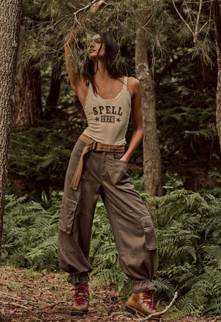 a-woman-stands-in-a-forest-wearing-a-beige-tank-top-with-spell-1976-printed-on-the-front-loose-brown-cargo-pants-a-tan-belt-and-red-hiking-boots