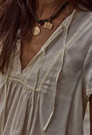 close-up-of-a-person-wearing-a-loose-short-sleeve-bohemian-style-top-in-a-light-beige-or-off-white-linen-like-fabric-with-lace-trim-and-tie-details-the-image-includes-part-of-the-persons-face-neck-and-hair-and-shows-a-dark-cord-necklace-with-wooden-pendants