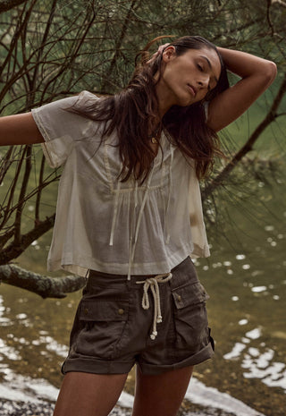 a-woman-posing-outdoors-near-water-and-trees-wearing-a-sheer-off-white-short-sleeve-tie-front-top-and-dark-olive-cargo-shorts