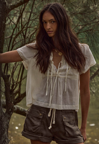a-woman-wearing-a-loose-white-short-sleeve-tie-front-blouse-and-brown-shorts-standing-outdoors-in-front-of-trees