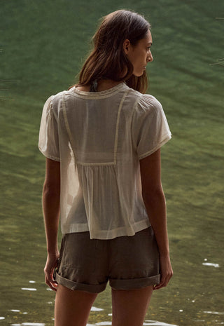 a-woman-viewed-from-behind-wearing-a-sheer-white-short-sleeve-blouse-and-olive-green-shorts-standing-outdoors-against-a-green-water-like-background