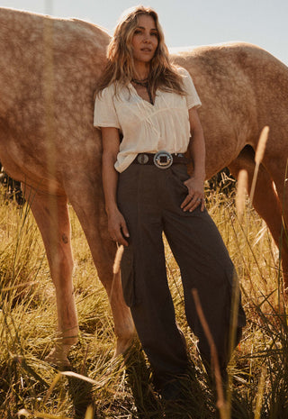 a-woman-stands-in-a-field-of-dry-grass-next-to-a-horse-wearing-a-cream-short-sleeve-blouse-dark-brown-wide-leg-cargo-pants-and-a-silver-belt