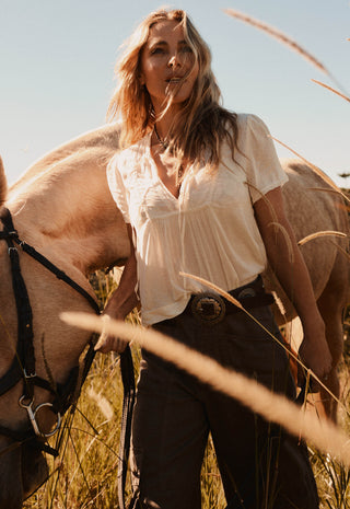 a-woman-standing-outdoors-in-a-field-with-a-horse-beside-her-wearing-a-loose-white-blouse-and-dark-pants-with-a-large-round-belt-buckle