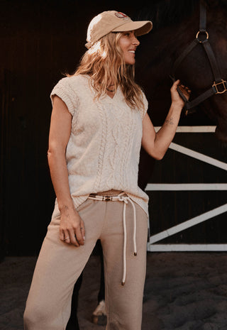 a-woman-wearing-a-beige-knit-sleeveless-top-matching-beige-drawstring-pants-and-a-beige-baseball-cap-stands-in-a-barn-holding-a-horses-bridle-the-background-shows-a-dark-barn-interior-with-white-painted-structural-lines
