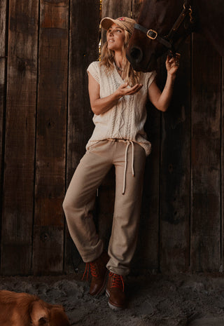 a-woman-posing-in-a-rustic-barn-setting-with-a-horse-wearing-a-cream-knit-sleeveless-top-beige-drawstring-sweatpants-and-brown-boots