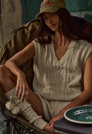 a-woman-wearing-a-beige-cable-knit-sleeveless-sweater-vest-and-matching-knit-socks-sits-in-a-camp-chair-wearing-a-khaki-baseball-cap-a-small-table-in-the-foreground-holds-a-green-plate