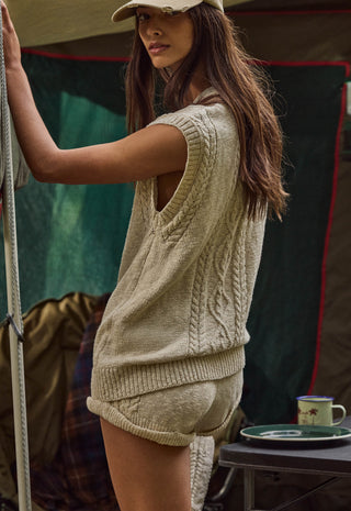 a-woman-wearing-a-beige-cable-knit-sleeveless-sweater-vest-and-matching-knit-shorts-standing-in-front-of-a-tent-with-a-camping-mug-on-a-table-in-the-background