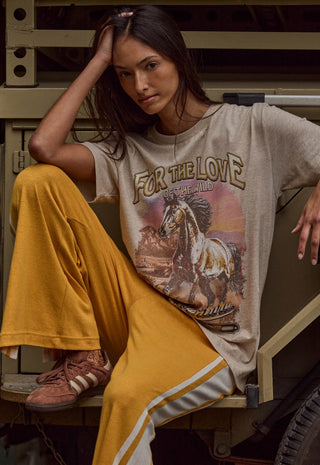 a-woman-models-a-beige-graphic-t-shirt-with-a-western-horse-design-and-the-text-for-the-love-of-the-wild-paired-with-mustard-yellow-pants-and-brown-sneakers-posing-on-a-rustic-wooden-bench