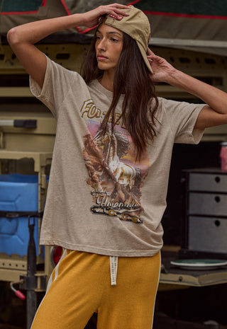 a-woman-wearing-a-beige-graphic-t-shirt-with-a-cowboy-on-the-front-paired-with-mustard-yellow-sweatpants-and-a-khaki-cap-posing-in-front-of-a-rustic-vehicle