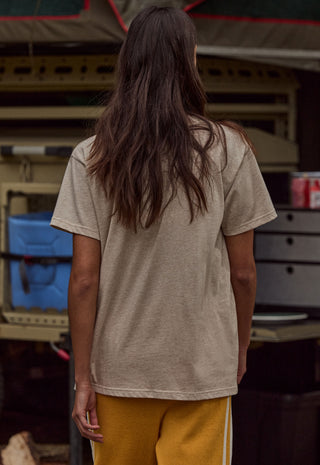 a-person-viewed-from-behind-wearing-an-oversized-light-beige-short-sleeve-t-shirt-and-mustard-yellow-pants-standing-in-front-of-a-rustic-outdoor-market-stall
