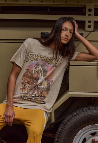 a-woman-wearing-a-beige-graphic-t-shirt-with-a-western-theme-and-yellow-pants-leaning-against-a-military-style-vehicle