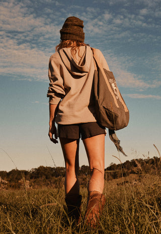 a-woman-seen-from-behind-walking-through-a-grassy-field-under-a-blue-sky-wearing-a-beige-hoodie-dark-shorts-a-brown-knit-beanie-and-carrying-a-large-brown-backpack