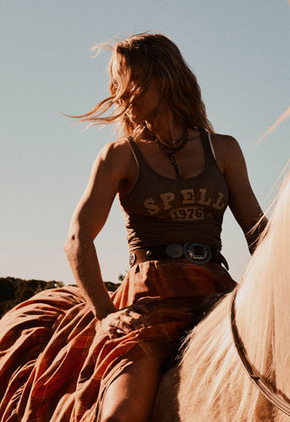 a-woman-riding-a-horse-outdoors-at-sunset-wearing-a-brown-tank-top-with-spell-1976-printed-on-the-front-a-rust-orange-plaid-skirt-and-a-large-silver-belt-buckle