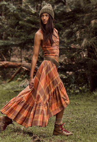 a-woman-wearing-a-rust-orange-and-brown-plaid-tiered-maxi-skirt-with-a-matching-cropped-top-a-green-knit-beanie-and-brown-lace-up-boots-standing-in-a-grassy-forest-clearing