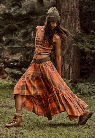 a-woman-wearing-a-rust-orange-and-brown-plaid-tiered-maxi-dress-with-a-matching-belt-a-knit-beanie-and-lace-up-boots-captured-mid-motion-while-walking-in-a-forest