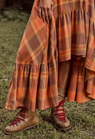 a-close-up-outdoor-shot-of-a-person-wearing-a-long-tiered-plaid-skirt-in-rust-orange-brown-and-yellow-tones-paired-with-brown-lace-up-boots-on-grass