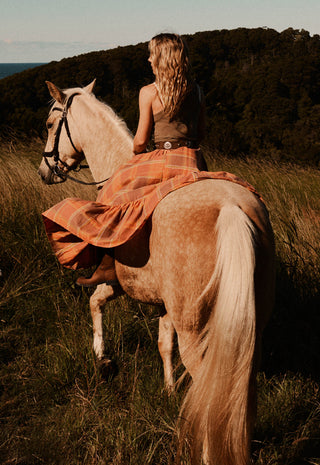 a-woman-riding-a-light-colored-horse-through-a-grassy-outdoor-landscape-at-golden-hour-wearing-a-brown-top-and-a-flowing-orange-plaid-skirt