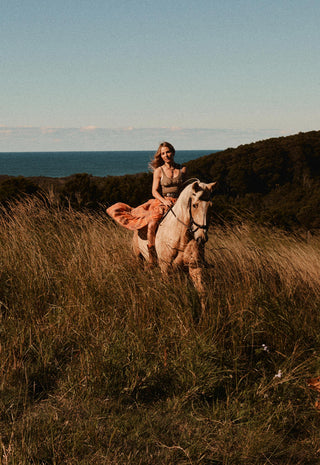 a-woman-rides-a-light-colored-horse-through-tall-dry-grass-on-a-hillside-with-ocean-and-dark-shrubbery-in-the-background-under-a-pale-blue-sky