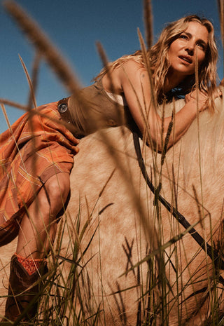 a-woman-in-a-desert-landscape-posing-on-a-light-colored-dune-with-tall-grass-in-the-foreground-she-is-wearing-a-sleeveless-beige-top-an-orange-plaid-skirt-and-brown-boots