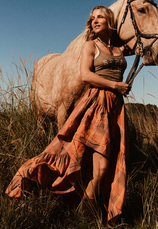 a-woman-in-a-rust-orange-plaid-maxi-dress-stands-outdoors-in-tall-grass-beside-a-light-colored-horse-holding-its-reins-under-warm-golden-sunlight