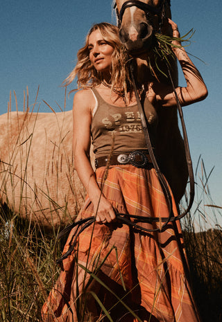 a-woman-in-a-rustic-western-style-outfit-stands-in-a-grassy-field-with-a-horse-beside-her-she-wears-a-brown-tank-top-with-western-text-graphics-and-a-long-rust-orange-plaid-skirt-holding-horse-reins-while-the-horse-looks-toward-the-camera-under-a-clear-blue-sky