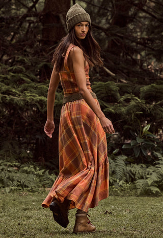 a-woman-wearing-a-sleeveless-orange-and-brown-plaid-maxi-dress-with-a-matching-beanie-and-brown-boots-standing-in-a-grassy-outdoor-setting-with-trees-in-the-background