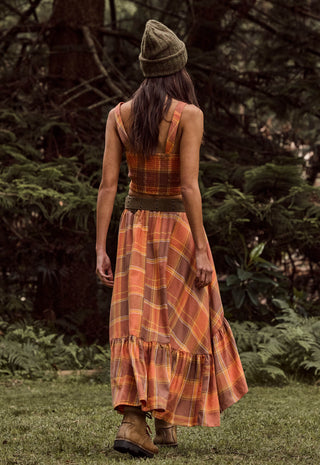 a-woman-viewed-from-behind-wearing-a-sleeveless-orange-and-brown-plaid-maxi-dress-with-a-matching-belt-and-a-knit-beanie-standing-on-grass-in-a-wooded-outdoor-setting