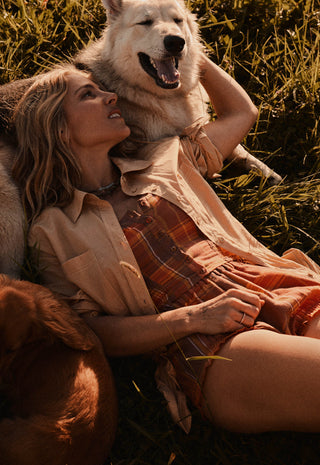 a-woman-lying-in-grass-outdoors-with-a-large-white-dog-behind-her-and-a-brown-dog-at-her-side-wearing-a-beige-shirt-and-rust-orange-plaid-shorts-or-dress-warm-golden-sunlight-illuminates-the-scene