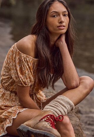 a-woman-crouching-outdoors-near-water-wearing-a-loose-off-the-shoulder-floral-dress-in-warm-tan-and-brown-tones-chunky-knit-socks-and-hiking-boots-with-red-laces