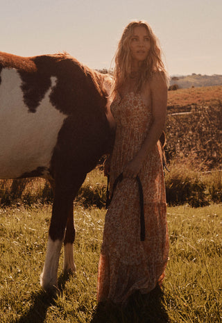 a-woman-in-a-sleeveless-patterned-maxi-dress-stands-in-a-grassy-field-next-to-a-brown-and-white-horse-with-a-warm-golden-sunset-background