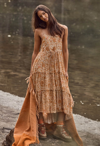 a-woman-stands-outdoors-by-a-body-of-water-wearing-a-sleeveless-tiered-maxi-dress-with-an-allover-floral-pattern-in-warm-tan-rust-and-cream-tones-the-dress-has-thin-straps-and-a-fringed-hem-and-she-is-also-wearing-an-orange-brown-fringed-throw-blanket-draped-over-one-side
