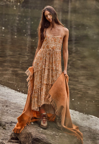a-woman-stands-outdoors-by-a-body-of-water-wearing-a-sleeveless-tiered-maxi-dress-with-an-abstract-print-in-warm-tan-beige-and-brown-tones-she-is-holding-a-large-rust-orange-fringed-throw-blanket-draped-over-the-ground