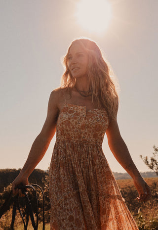 a-woman-outdoors-in-warm-golden-hour-sunlight-wearing-a-strapless-floral-maxi-dress-holding-a-dark-woven-strap-in-one-hand-the-background-is-a-bright-sunlit-sky-with-blurred-brush-and-hills