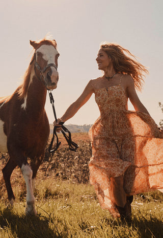 a-woman-in-a-flowing-peach-floral-dress-walks-beside-a-brown-and-white-horse-in-an-open-field-holding-the-horses-reins-under-warm-golden-sunlight