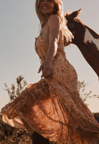 a-woman-wearing-a-flowing-rust-orange-patterned-maxi-dress-is-captured-outdoors-with-a-brown-and-white-horse-beside-her-against-a-clear-sky