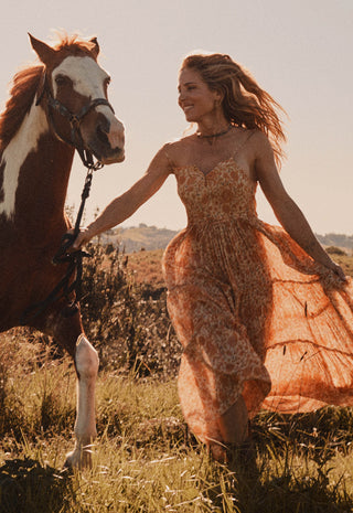 a-woman-in-a-flowing-rust-orange-boho-dress-walks-beside-a-brown-and-white-horse-in-an-outdoor-grassy-field-at-golden-hour