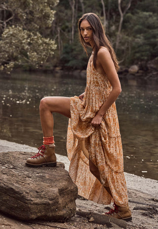 a-woman-stands-on-a-large-rock-beside-a-river-wearing-a-sleeveless-tiered-maxi-dress-in-a-warm-tan-and-orange-floral-print-paired-with-brown-lace-up-boots