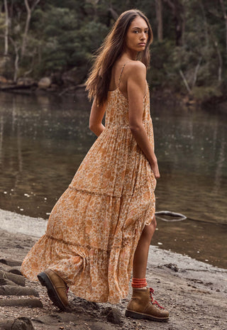 a-woman-wearing-a-flowing-orange-and-beige-floral-maxi-dress-with-brown-hiking-boots-stands-on-a-rocky-riverside-path-with-water-and-trees-in-the-background