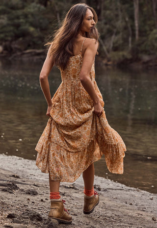 a-woman-wearing-a-rust-orange-and-beige-floral-print-midi-dress-standing-on-a-rocky-riverbank-with-water-and-trees-in-the-background