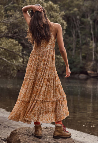 a-woman-stands-on-a-log-by-a-river-wearing-a-sleeveless-tiered-maxi-dress-with-an-orange-and-brown-floral-pattern-she-is-shown-from-behind-looking-out-over-the-water-with-trees-in-the-background