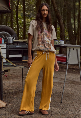 a-woman-stands-outdoors-in-a-wooded-campsite-setting-wearing-loose-mustard-yellow-drawstring-pants-with-white-side-stripes-a-beige-graphic-t-shirt-and-brown-shoes