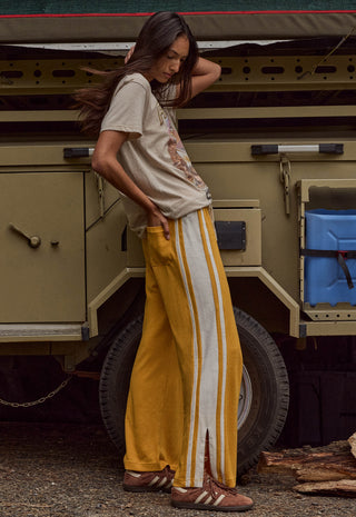 a-woman-stands-outdoors-in-front-of-a-military-style-vehicle-wearing-a-beige-graphic-t-shirt-mustard-yellow-track-pants-with-white-side-stripes-and-brown-sandals