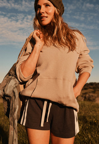 a-woman-outdoors-wearing-a-beige-oversized-sweatshirt-dark-brown-shorts-with-white-side-stripes-and-a-dark-knit-beanie-with-a-camouflage-jacket-draped-over-her-arm-against-a-blue-sky-background